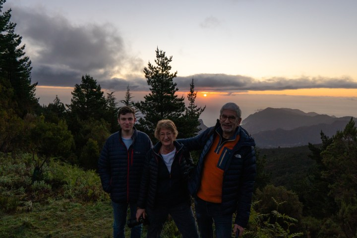 Three people in jackets standing in front of a sunset view with trees and mountains.