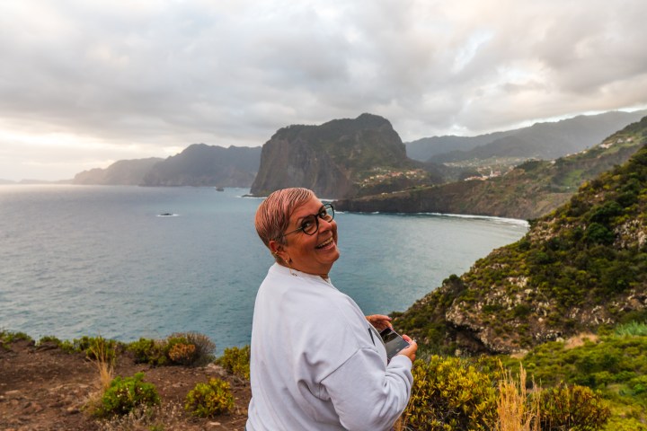 Person smiling at ocean cliff view with cloudy sky and green mountains.
