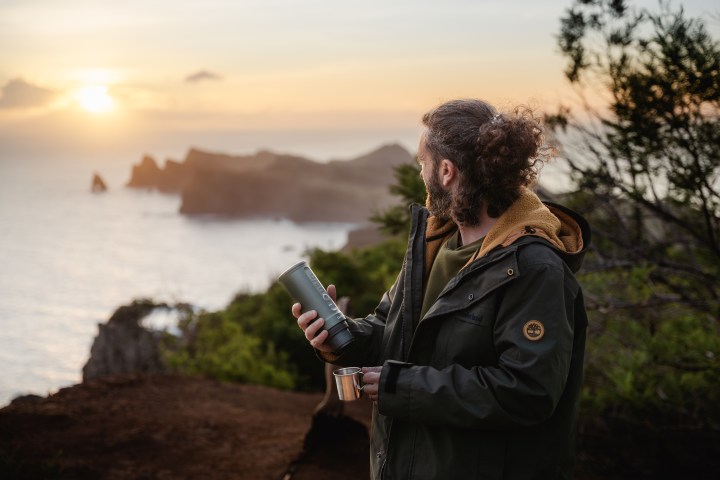 Person with a thermos overlooks ocean and cliffs at sunset.